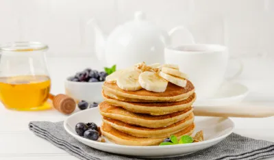 A stack of oatmeal banana pancakes with slices of fresh bananas, walnuts and honey on top with cup of tea on a white wooden background. A healthy breakfast. Copy space