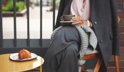 Woman sitting in outdoor cafe with cup of cappuccino coffee in her hands and croissant on table comfortably wrapped in gray plaid scarf, Because it became chilly.
