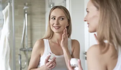 Young caucasian woman smiling and applying face cream in the mirror reflection